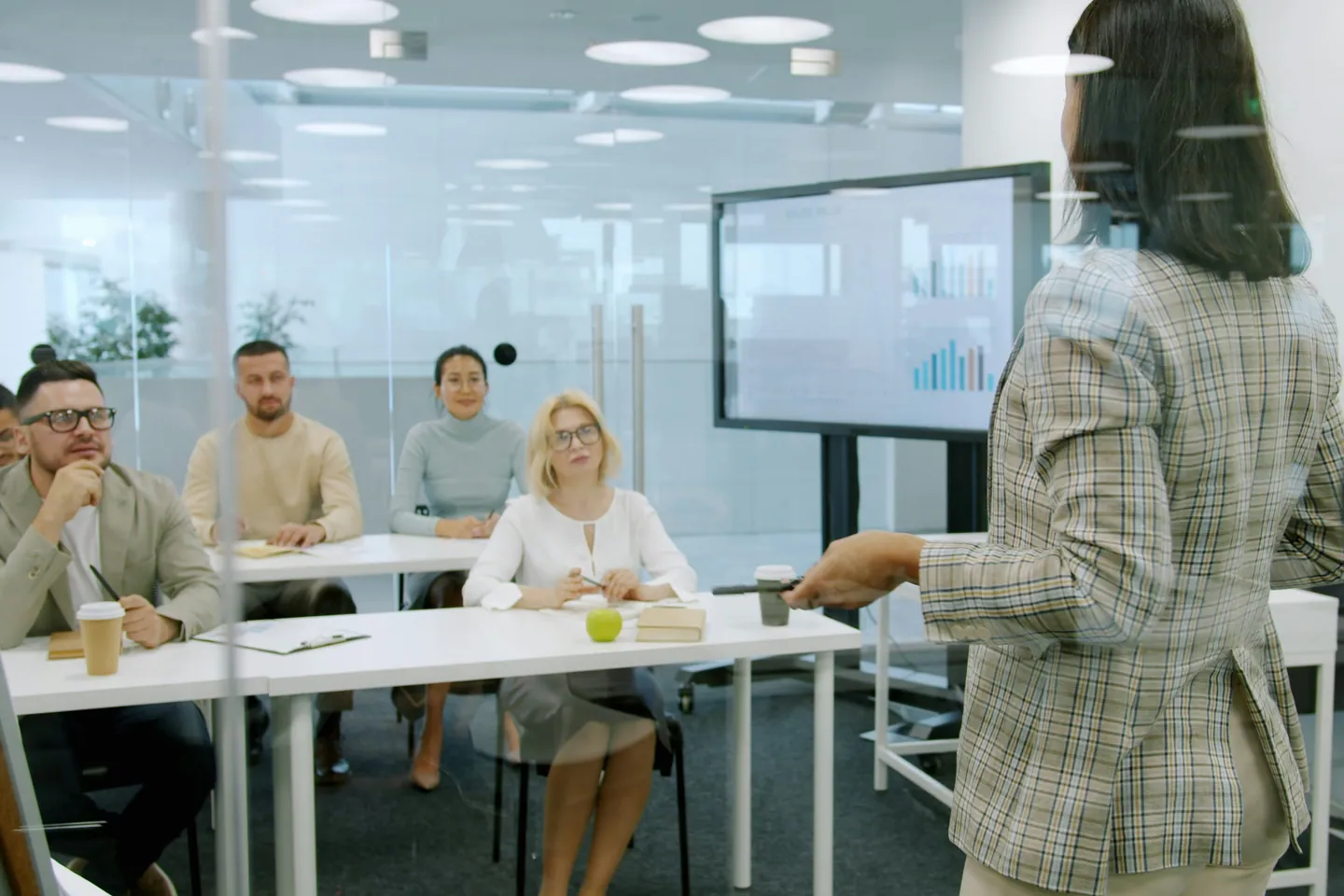 Formatrice devant un écran de graphiques, face à un petit groupe de salariés assis à des tables dans une salle de réunion vitrée